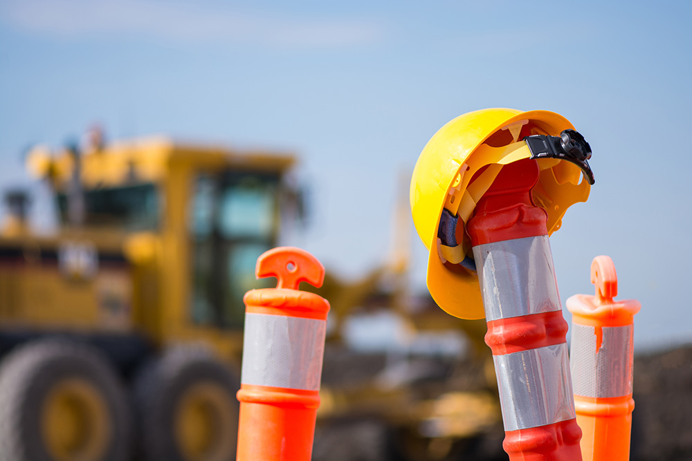 A hardhat sits on a road cone with a bulldozer in the background.