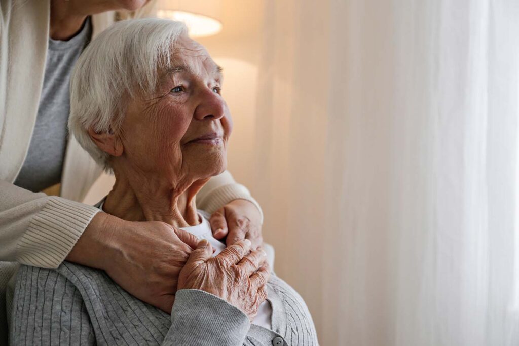 An elderly woman sits with a younger woman putting her arms around her.